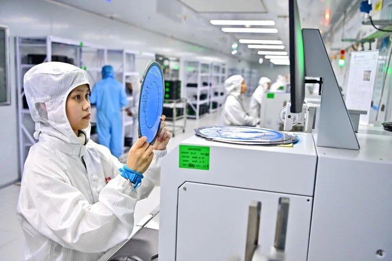 A technician in white overalls holds up a blue disc shaped semiconductor whist work in a semiconductor factory in China