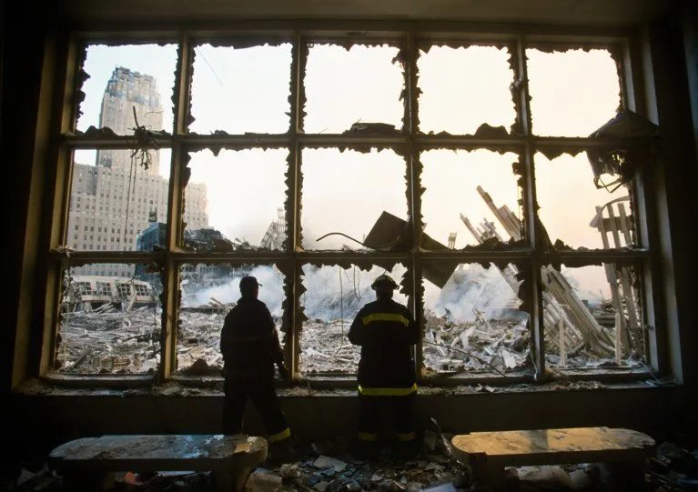 Two figures sihouetted against a large window with smashed glass panes looking out onto the smouldering rubble of the World Trace Center following the 911 terror attacks