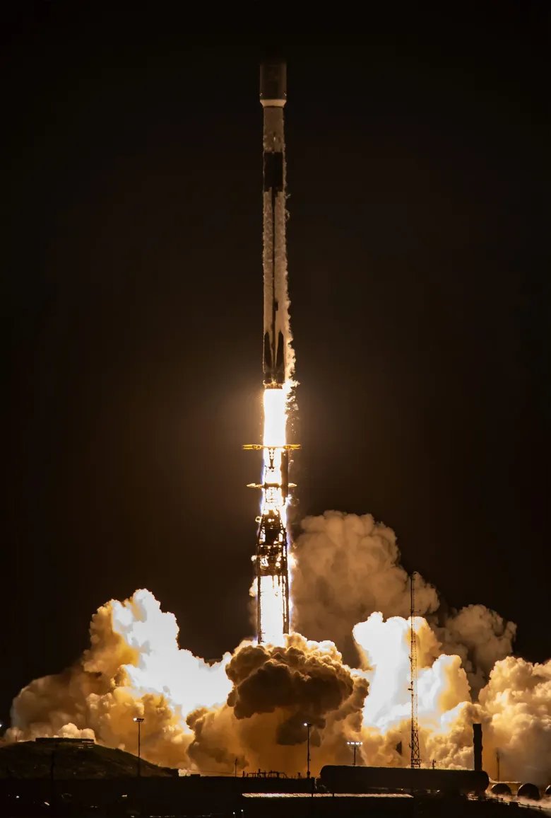 a white and black rocket lifts off into the night sky its engine plume glowing orange-white lights its launch pad