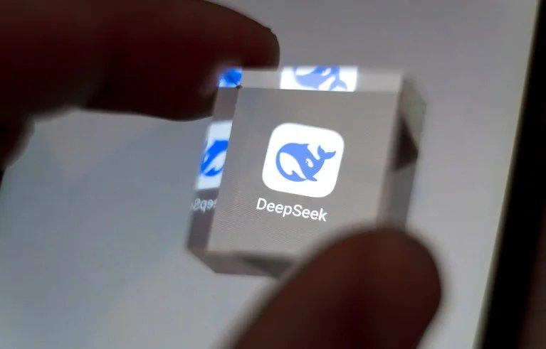 The DeepSeek application icon displayed on a smartphone screen is magnified by a clear cube held between thumb and forefinger